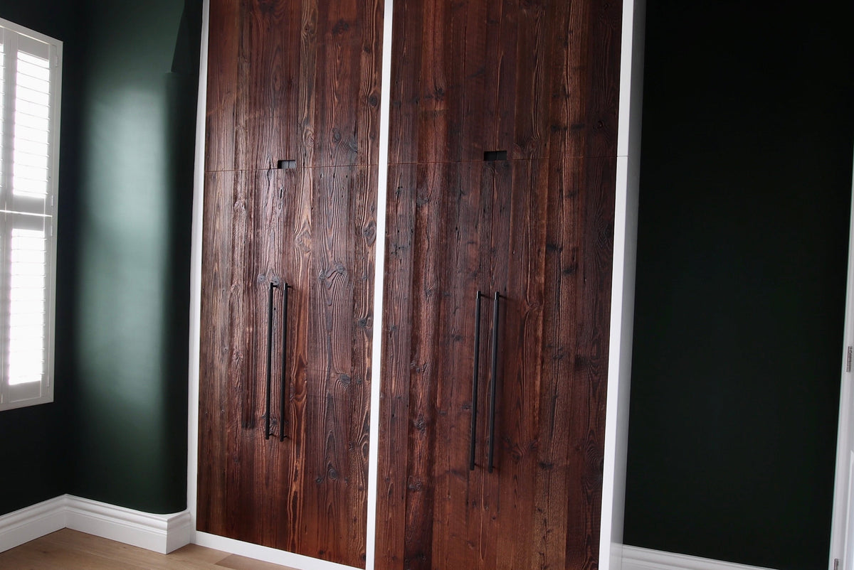 A large built-in wardrobe with two dark brown wood doors and vertical black handles, set into a room with dark green walls and light wood flooring.