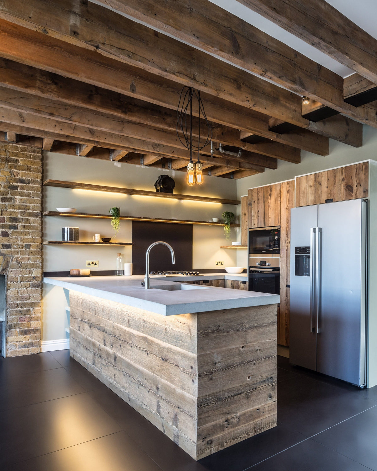 A modern open-plan kitchen with a large reclaimed solid wood-block island, reclaimed wood cabinet doors and shelves, exposed wooden ceiling beams, a brick fireplace, and a staircase on the right.