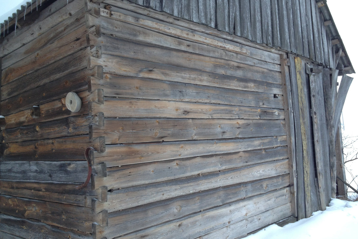 A close-up of an old weathered wooden structure with horizontal log siding, a dark wood roof, and snow on the ground. Cladding 