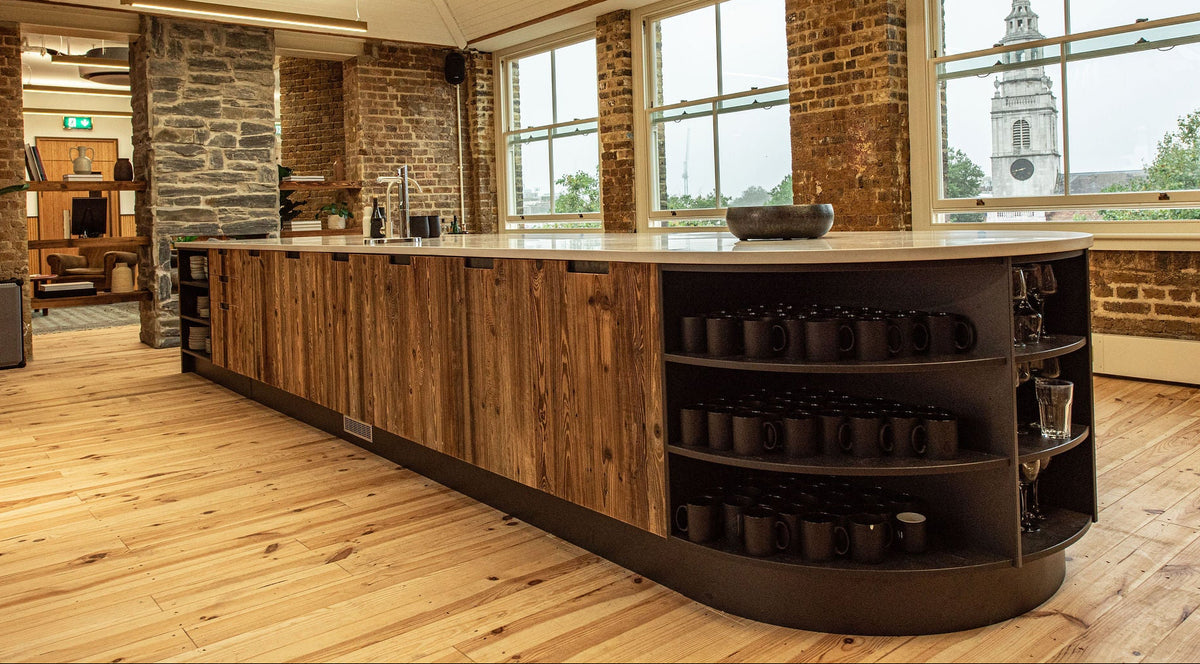 A modern kitchen island with rustic wood cabinets, a light stone countertop, and built-in black shelving filled with mugs, set in a loft-style room with exposed brick walls and large windows.
