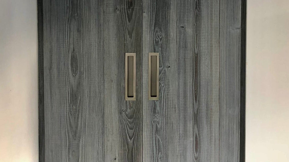 A tall double-door wardrobe or closet with dark gray woodgrain doors and slim vertical gold handles, set against off-white walls.