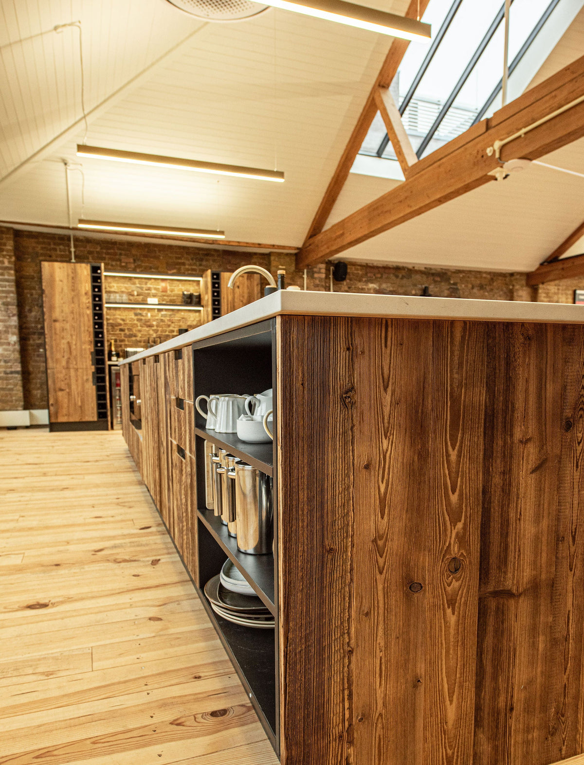 A long kitchen island with richly stained brown wood paneling and a light countertop, set in a loft-style kitchen with exposed brick walls, wooden beams, and a skylight.