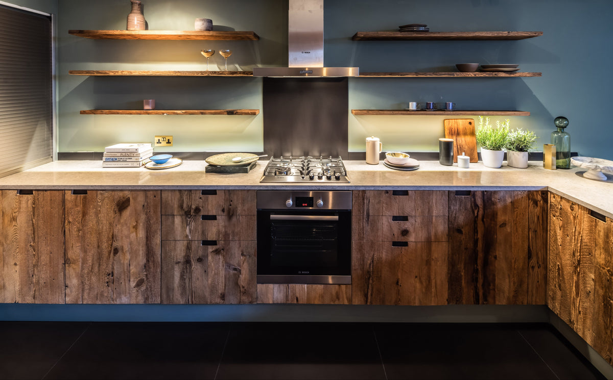 A modern kitchen island with rustic dark wood cabinets, a stainless steel gas range, built-in oven, and a central stainless steel range hood. The background wall is a muted blue-green, decorated with floating wooden shelves holding ceramic vases and dishes.