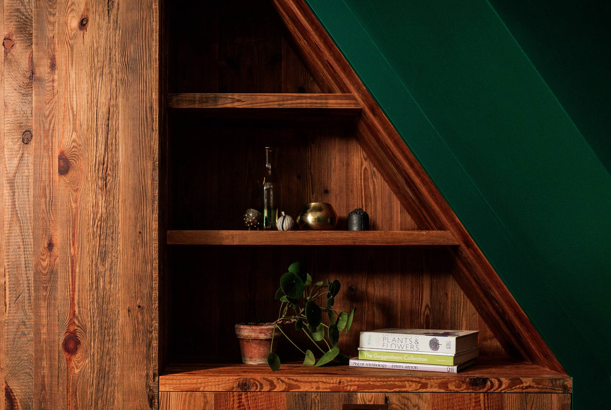 A rustic wooden slat cabinet with integrated shelves, styled with books and a small potted plant against a dark green slanted wall.