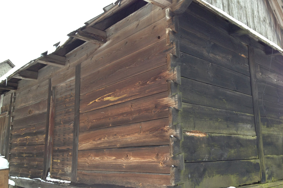 Close-up of the corner of a rustic wooden building with dark stained logs and snow patches at the base. Dovetail corners and chunky solid wood and door, cladding