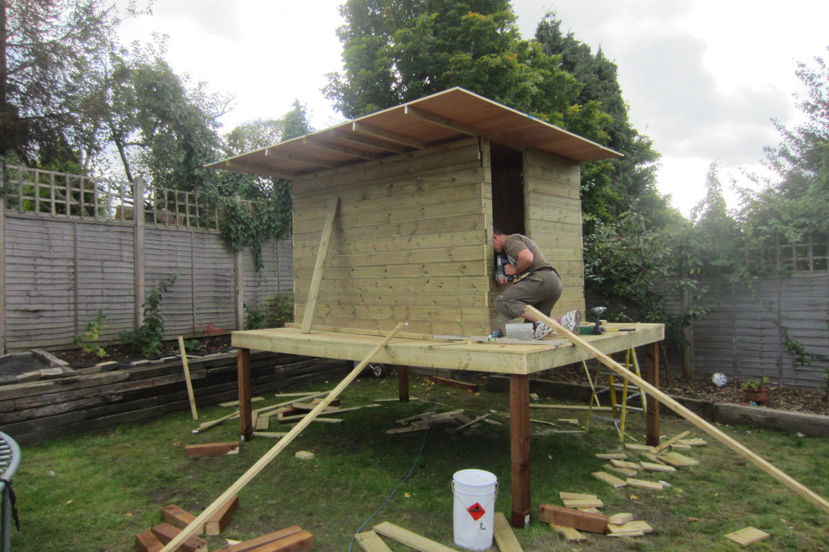 A man building a wooden garden shed on a raised platform in a backyard. The shed features light-colored wooden planks on the walls and a darker brown frame, supported by a raised wooden deck with legs.