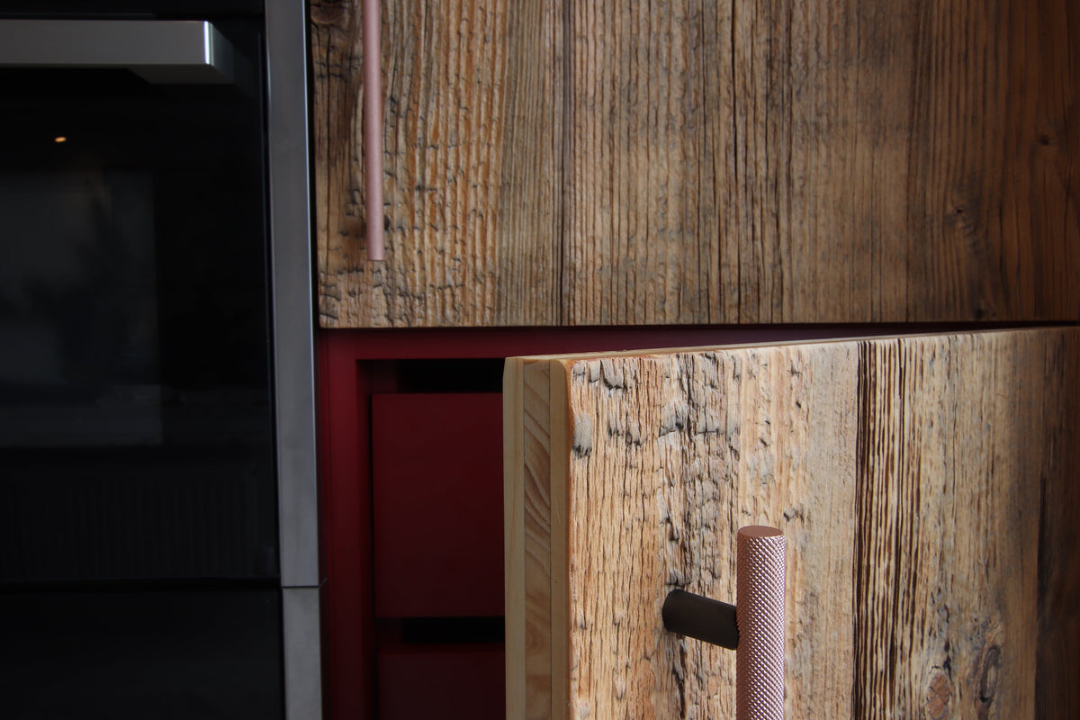 Close-up of reclaimed wood cabinetry, in kitchen with copper handle and a red drawer front and a dark stainless steel appliance edge on the right.