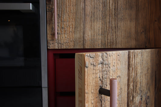 Close-up of reclaimed wood cabinetry, in kitchen with copper handle and a red drawer front and a dark stainless steel appliance edge on the right.