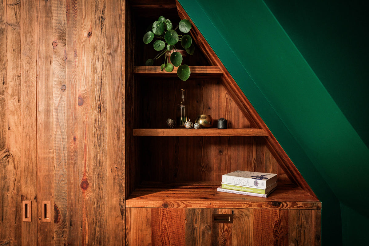 Bespoke fitted shelving, worktop, vanity unit and cabinetry crafted from reclaimed barn wood, featuring warm natural textures, angled shelves, and brass details beside a deep green wall