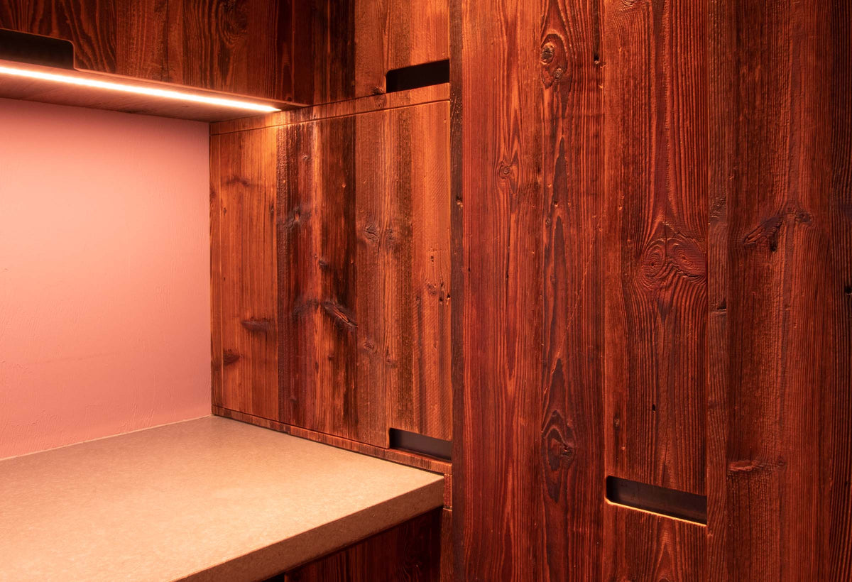 Close-up of reclaimed barn wood cabinetry with recessed handles and warm LED under-shelf lighting against a soft pink wall