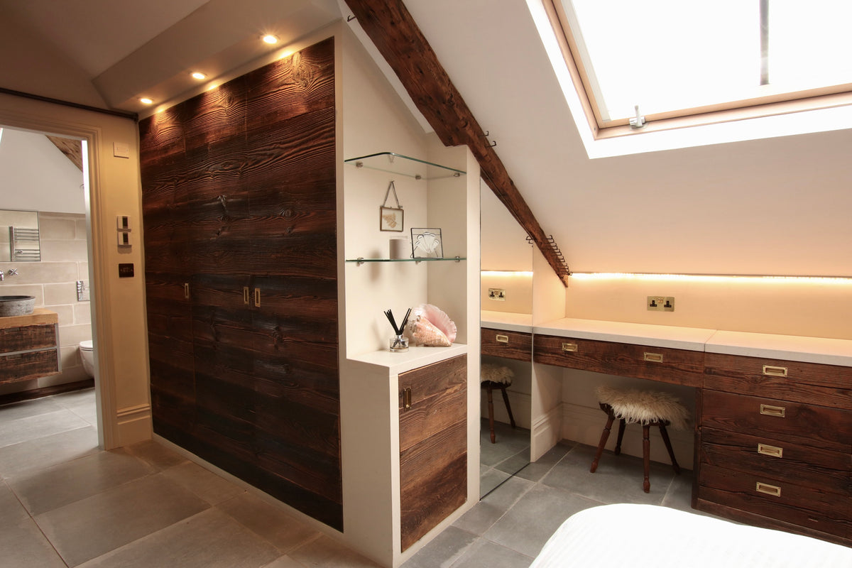 Bespoke fitted wardrobe and dressing area crafted from reclaimed barn wood in a loft bedroom, with skylight, glass shelving and warm integrated lighting