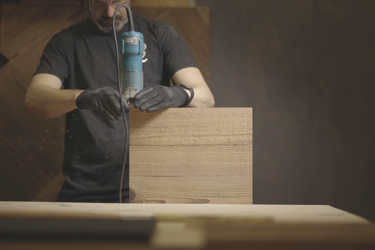 A man wearing a black t-shirt and black work gloves uses a blue and gray orbital sander to smooth a large wooden board in a workshop setting.