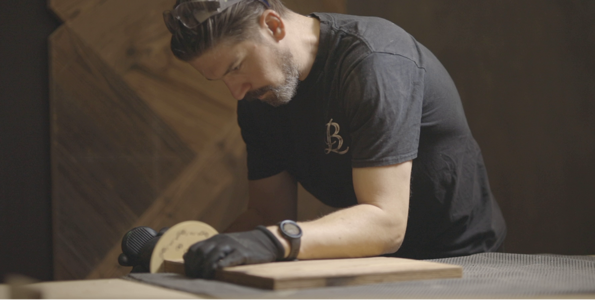 A man wearing a black t-shirt with a small gold monogram logo and black gloves works at a wooden table in a dimly lit workshop.