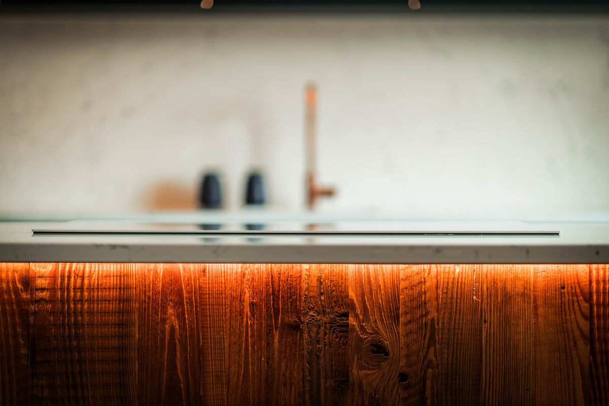 A close-up, low-angle view of a modern kitchen sink with a matte black basin, a warm copper faucet, and a prominent wood-look front panel illuminated by warm under-cabinet lighting against a light textured wall.